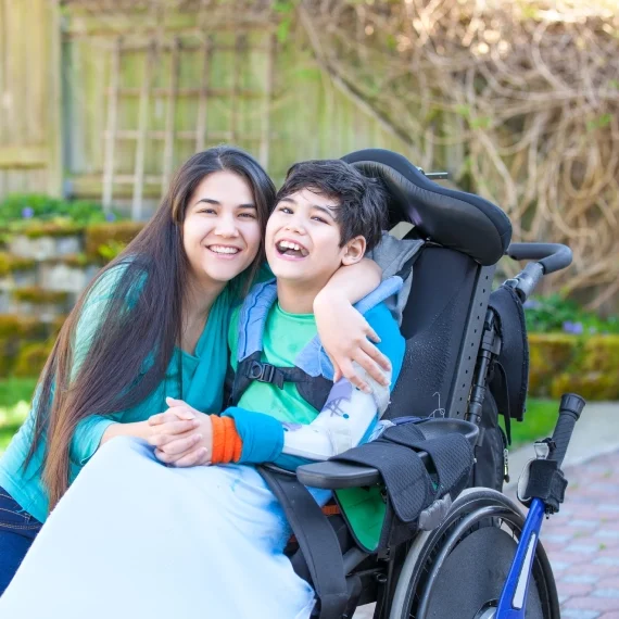 A woman smiles and hugs a boy sitting in a wheelchair outdoors. They are on a paved area with a garden wall and greenery in the background.