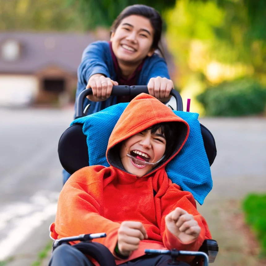A young person pushes another young person in a wheelchair outdoors. Both are smiling and enjoying the moment.