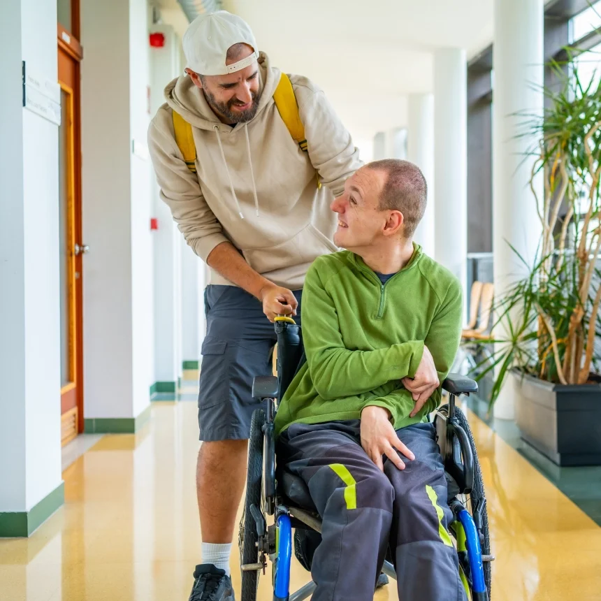 A man in a wheelchair smiles at another man pushing him through a bright hallway with plants.